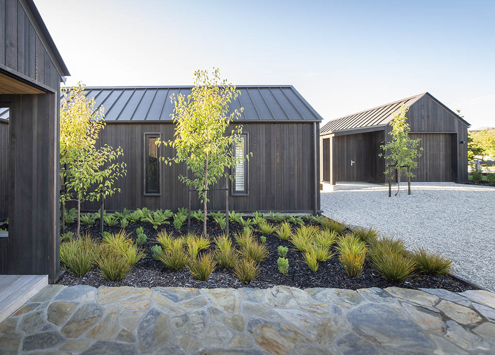 Sheltered arrival path between gables at Wānaka Barn House with schist paving, trees and grasses