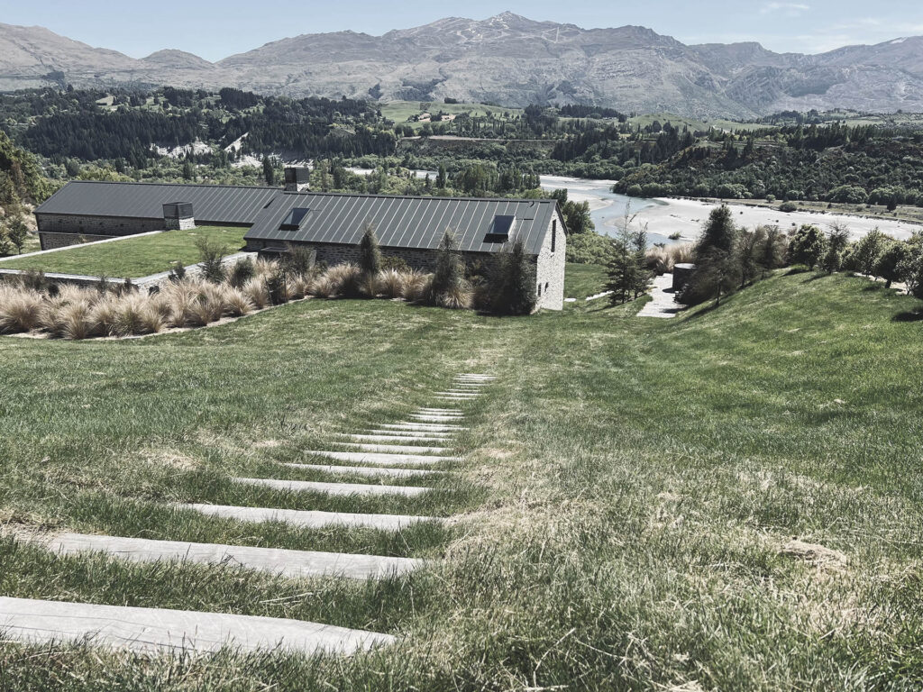 Sleeper steps down a sloped lawn at Shotover River House with the river and mountains beyond.