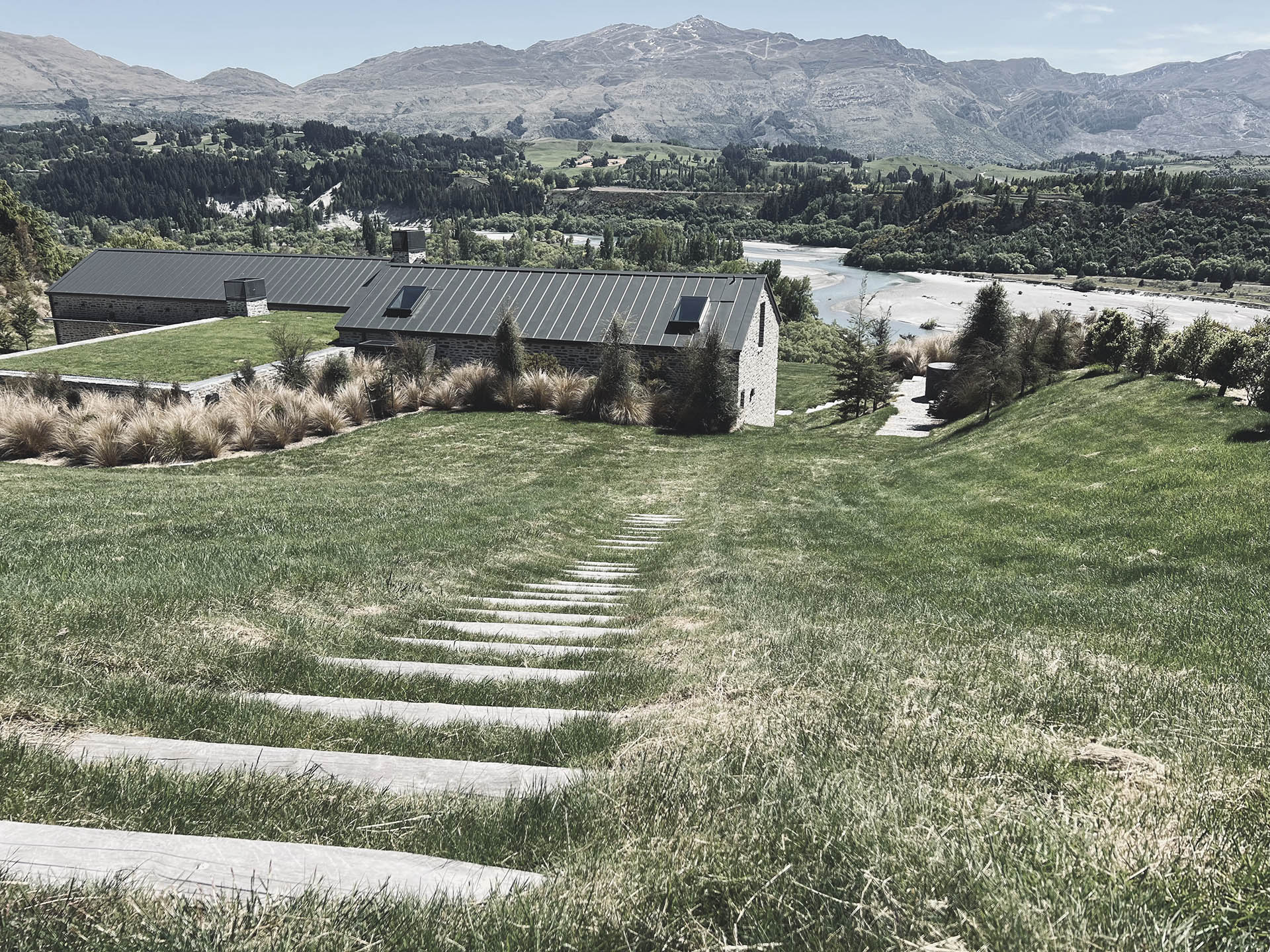 Sleeper steps down a sloped lawn at Shotover River House with the river and mountains beyond.
